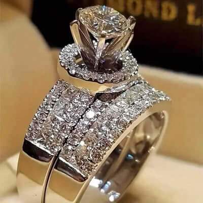 A close-up of a silver-toned engagement ring with a large round center stone and a matching band set with small clear stones, displayed on a beige ring box.
